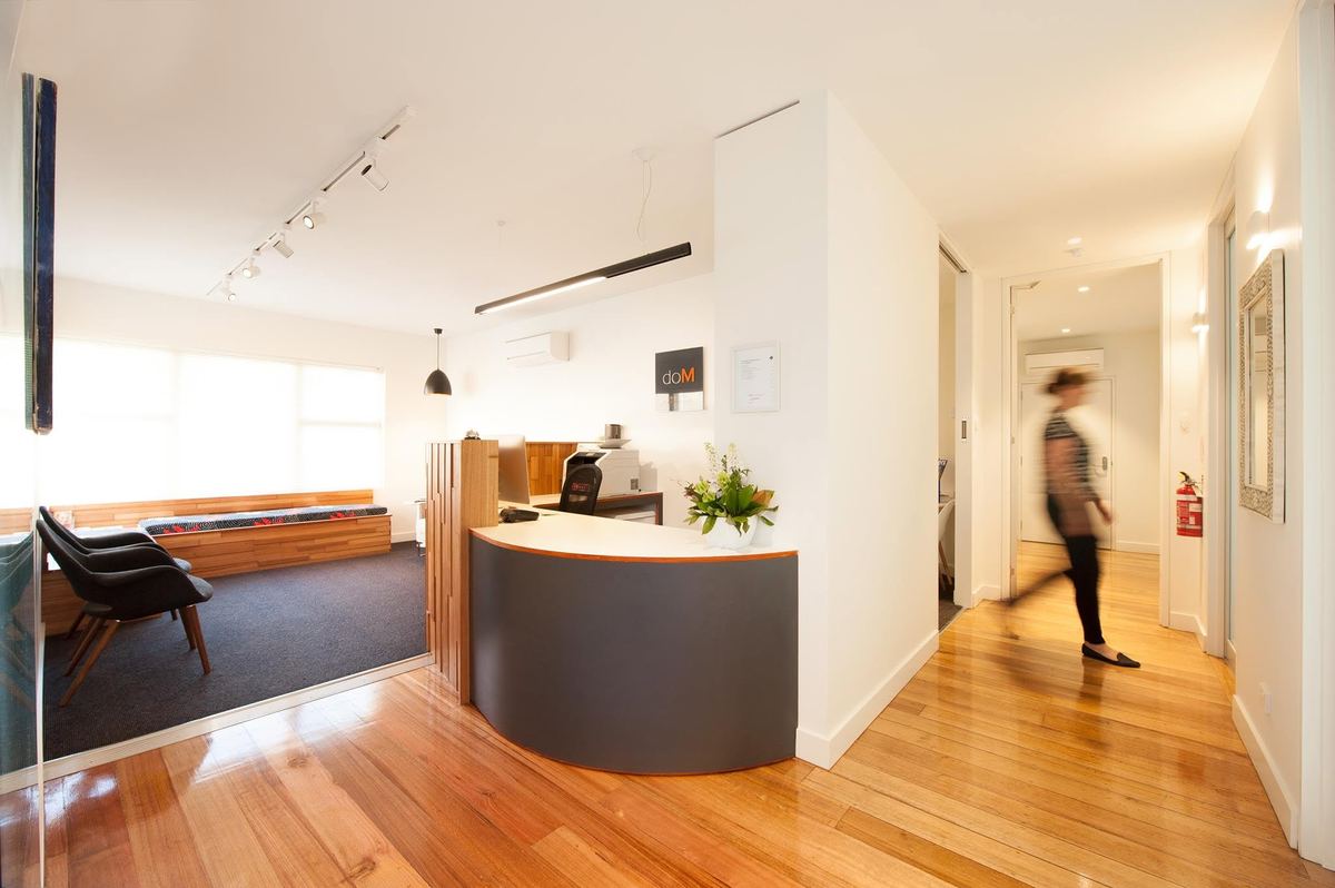 The doM reception: a curved charcoal front desk with orange trim, timber floors and a sunlit waiting area.